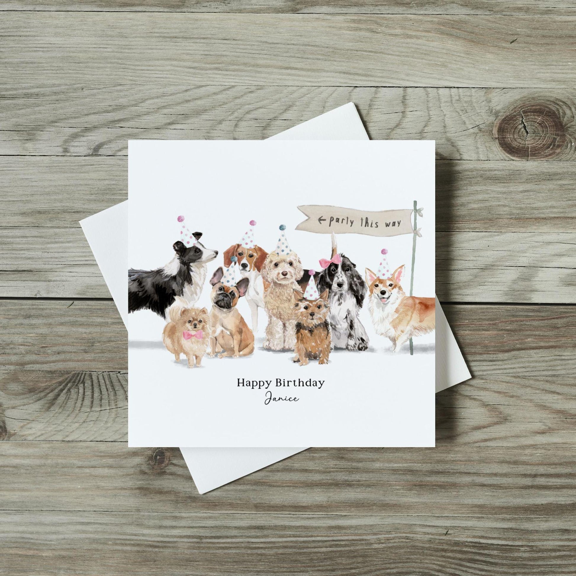 A square, white card with 8 dogs on and a sign saying&quot;party this way&quot;.  Text reading Happy Birthday Janice is written under the image.  Dog breeds include a cockerpoo, a border collie, a beagle, a cocker spaniel.